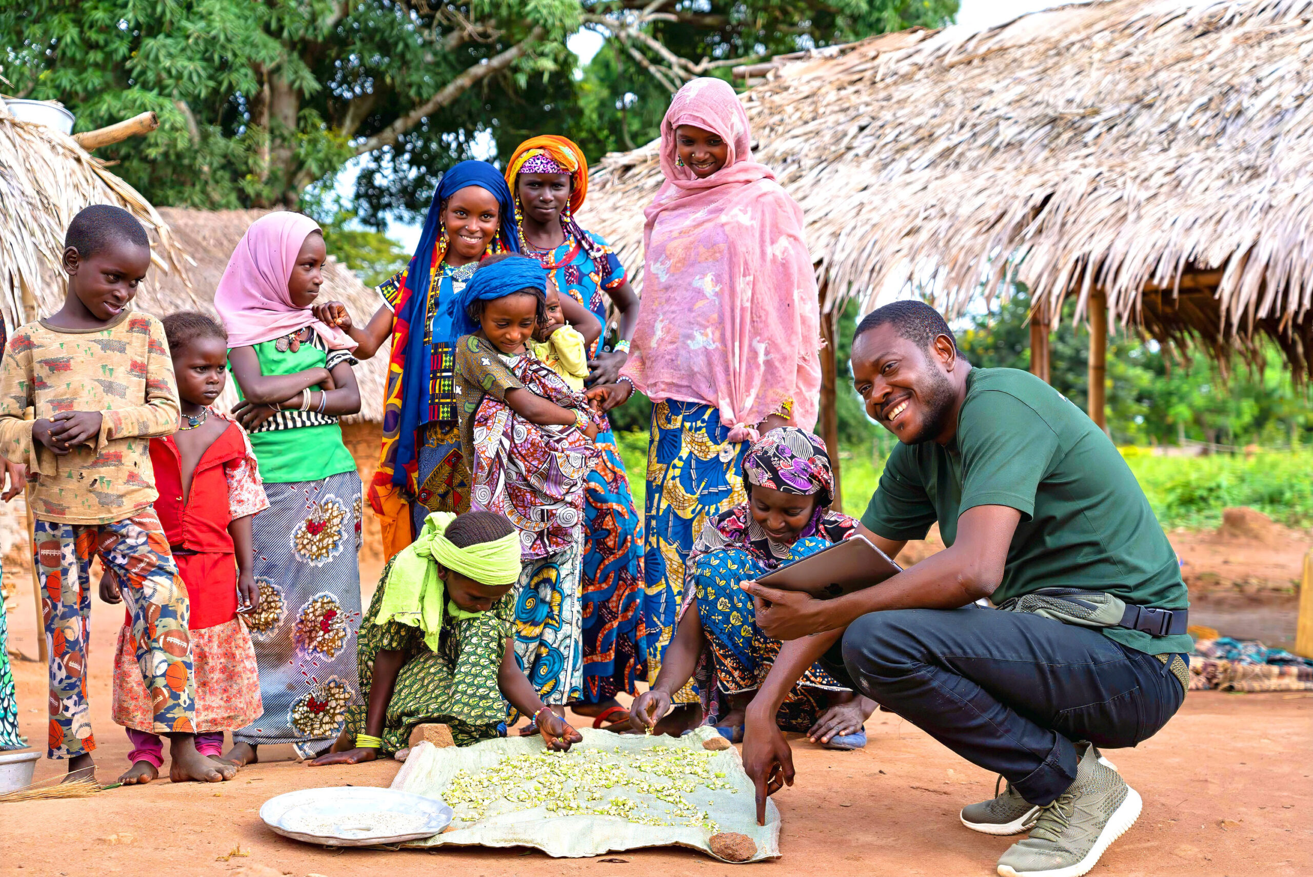 Women and children spread okra on a cloth in a rural village. Dodo Moke leans over the okra and smiles with them.